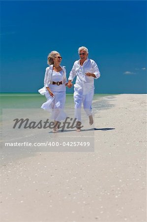 Happy senior man and woman couple running, laughing and holding hands on a deserted tropical beach with bright clear blue sky