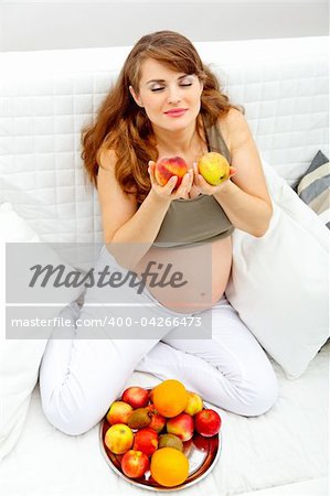 Beautiful pregnant woman sitting on sofa at home and enjoying fruits
