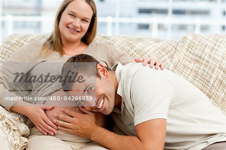 Smiling future dad listening to the belly of his wife sitting on the sofa in the living room