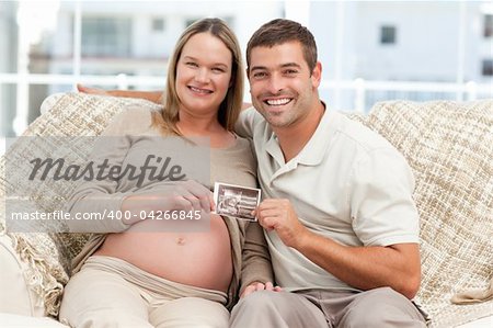 Cheerful couple of future parents holding an echography sitting on the sofa