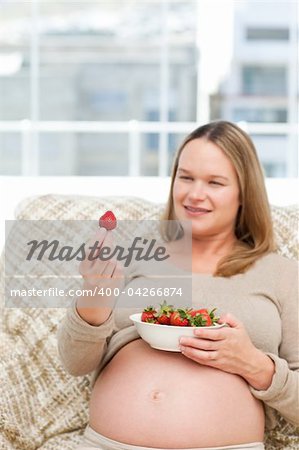 Adorable pregnant woman looking at a strawberry while relaxing on the sofa