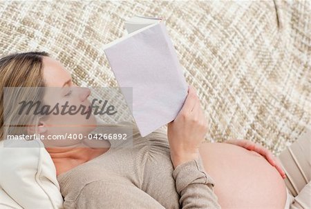 Cute pregnant woman reading a book lying on the couch at home