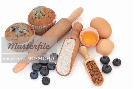 Blueberry muffin baking ingredients including, blueberries, muffins, eggs, wholegrain flour, brown sugar and wooden kitchen utensils, over white background.