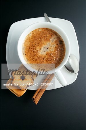 White porcelain cup of freshly brewed coffee top view close-up arranged with two sandwich-biscuits, cinnamon stick, spoon and plate on dark background