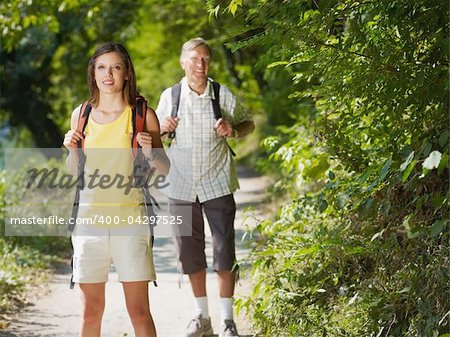 senior man and young woman walking with backpack in wood, looking at camera and smiling. Horizontal shape, front view, copy space