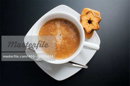 White porcelain cup of freshly brewed coffee top view close-up arranged with two sandwich-biscuits, spoon and plate on dark background