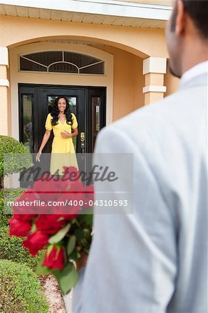 A romantic African American man bringing flowers to his happy wife or girlfriend who is standing waiting for him at the door of their home.