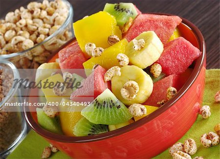 Fresh fruit salad made of banana, kiwi, watermelon and mango pieces in orange bowl with cereals (puffed wheat and puffed chocolate quinoa) (Selective Focus, Focus on the front of the bowl and the fruits in the front)