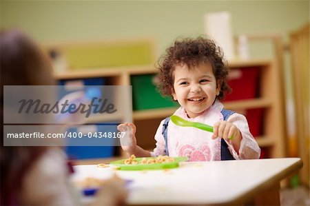 Caucasian and hispanic female preschoolers eating pasta and smiling. Horizontal shape, waist up, focus on background