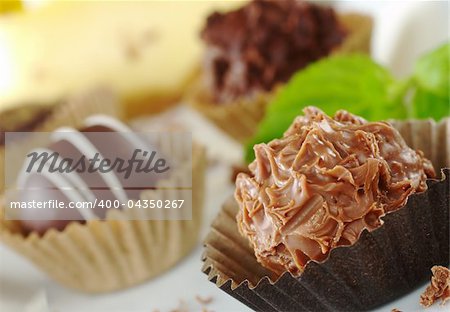 Chocolate truffles with mint leaf, fine chocolate shavings and a yellow cake in the background (Very Shallow Depth of Field, Focus on the front of the first praline)