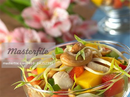 Fresh Asian salad with chicken, mango, cucumber, bean sprouts, red bell pepper and peanuts in glass bowl with inca lily in the back (Selective Focus, Focus on the cashew nut and the bean sprout on the top)
