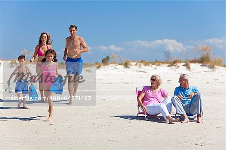 A happy family of mother, father, two children, son and daughter, running having fun on a sunny beach while the grandparents sit looking on and laughing.