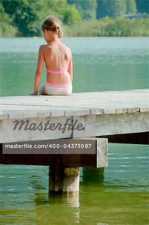 Cute little girl sitting on a pier and looking at the water. Vertical view