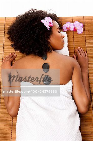 Beautiful happy relaxed Latina-African woman at health day spa with hot lastone therapy massage treatment laying in white towel on bamboo table decorated with orchids.