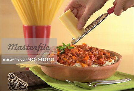 Grating cheese over spaghetti bolognaise with minced meat, carrot, tomato and onion, garnished with oregano leaf (Selective Focus, Focus on the oregano leaves on the spaghetti and the cheese held in the hand)