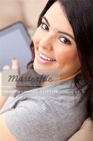 Beautiful happy young Latina Hispanic woman smiling and using a tablet computer or iPad at home on her sofa