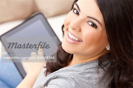 Beautiful happy young Latina Hispanic woman smiling and using a tablet computer or iPad at home on her sofa