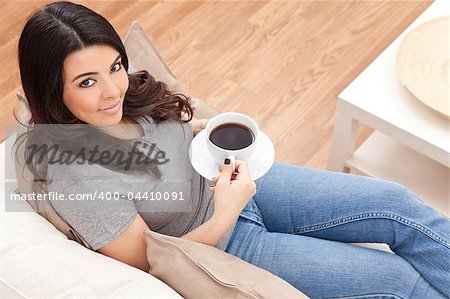 A beautiful young Latina Hispanic woman or girl with a wonderful enigmatic smile drinking tea or coffee from a white cup at home on her sofa