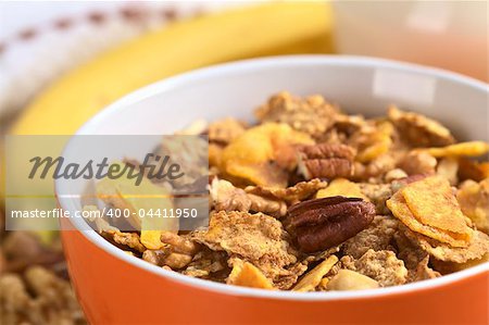 Healthy and delicious breakfast with a bowl full of wholewheat flakes mixed with banana chips, walnuts and pecan nuts with a fresh banana and milk in the back (Selective Focus, Focus on the pecan nut one third into the bowl)