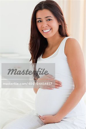 Portrait of a good looking pregnant woman touching her belly while sitting on a bed at home