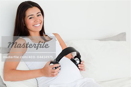 Frontal view of an attractive pregnant woman putting headphones on her belly while lying on a bed at home