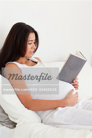 Charming pregnant woman reading a book while lying on a bed at home