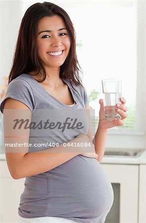 Beautiful pregnant woman holding a glass of water while standing in the kitchen
