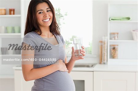Attractive pregnant woman holding a glass of water while standing in the kitchen