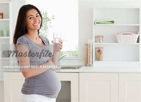 Good looking pregnant woman holding a glass of water while standing in the kitchen