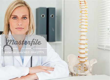 Serious blonde doctor with model spine next to her looks into camera in her office