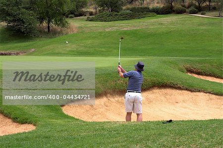 Golfer hitting the ball out of the sand bunker straight onto the green. The golf ball is flying in the air.