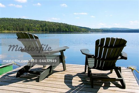 Two adirondack wooden chairs on dock facing a blue lake with clouds reflections