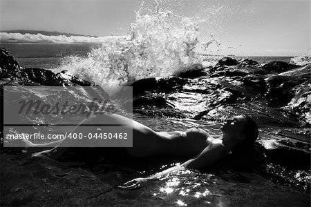 Caucasian young adult woman lying nude in tidal pool in Maui with wave crashing in distance.