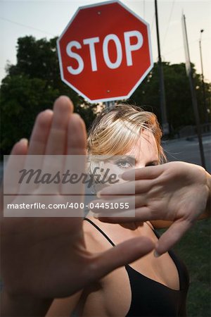 Caucasian mid-adult blonde woman holding hands up to camera in front of stop sign.