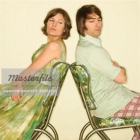 Caucasian mid-adult couple wearing vintage clothing sitting back to back in green vinyl chairs with arms crossed looking angry.