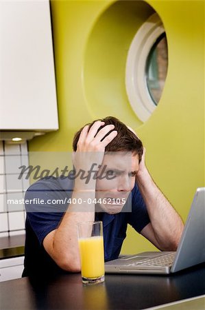 A worried computer user sits in his kitchen with a desperate look on his face