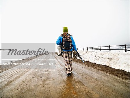 Young woman in winter clothes walking down muddy dirt road holding snowboard and boots.