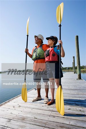African American middle-aged couple standing on boat dock holding paddles wearing life preservers.