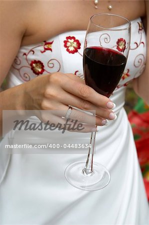 The hand of the bride in the white dress decorated by a red embroidery, holds a glass with red wine