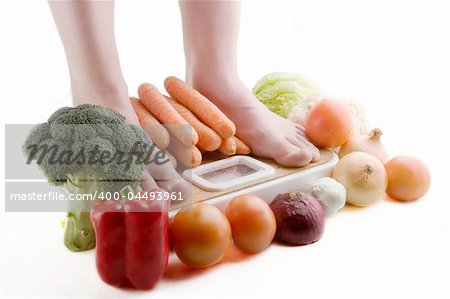 A pair of female feet standing on a bathroom scale with vegetables pilled around.