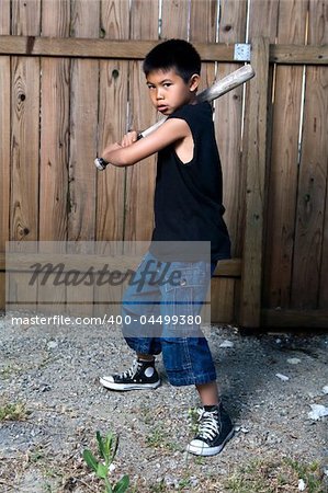 Young asian boy standing outside beside a tall wooden fence wearing jeans and black tshirt holding a baseball bat