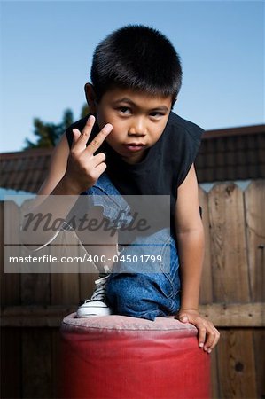 Young asian boy kneeling on top of a red punching bag outside beside a tall wooden fence wearing jeans and black tshirt making hand gesture