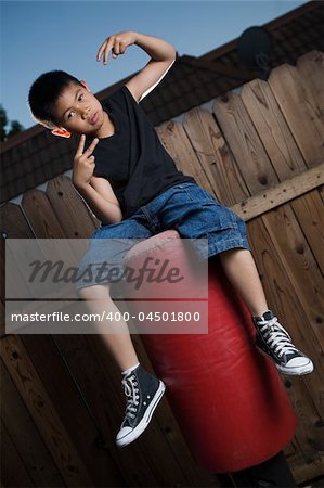 Young asian boy sitting on top of a punching bag outside beside a tall wooden fence smiling wearing jeans and black tshirt making hand signals