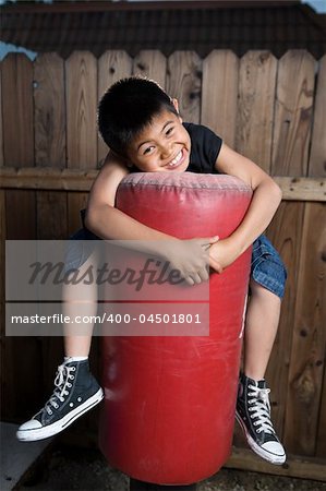 Young asian boy holding on to a punching bag outside beside a tall wooden fence smiling wearing jeans and black tshirt