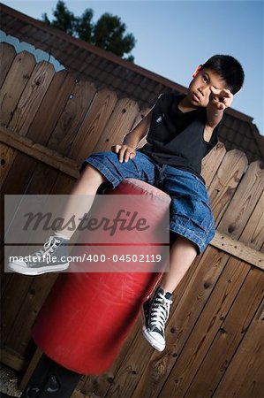 Young asian boy sitting on top of a punching bag outside beside a tall wooden fence smiling wearing jeans and black tshirt