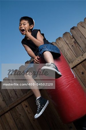 Young asian boy sitting on top of a punching bag outside beside a tall wooden fence resting his chin on hand wearing jeans and black tshirt