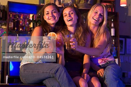 Three young women sitting on a bar counter, toasting the camera