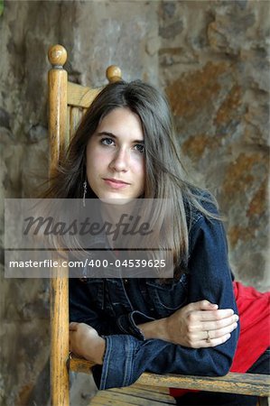 Female teen sits in a rocking chair on stone porch.  Denim jacket and red shirt. Solemn expression.