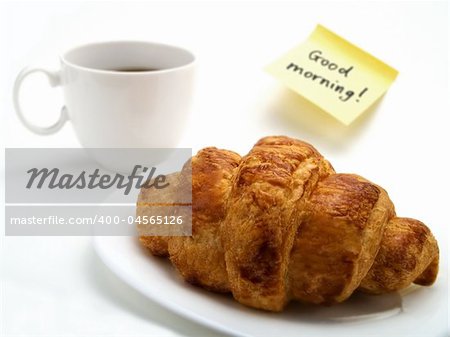 A crispy croissant in a white plate, a cup of fresh coffee and a yellow note with this text: 'Good morning!'