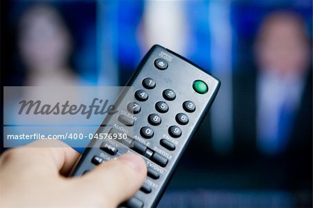Hand holding a remote control pointing to a blurred TV program